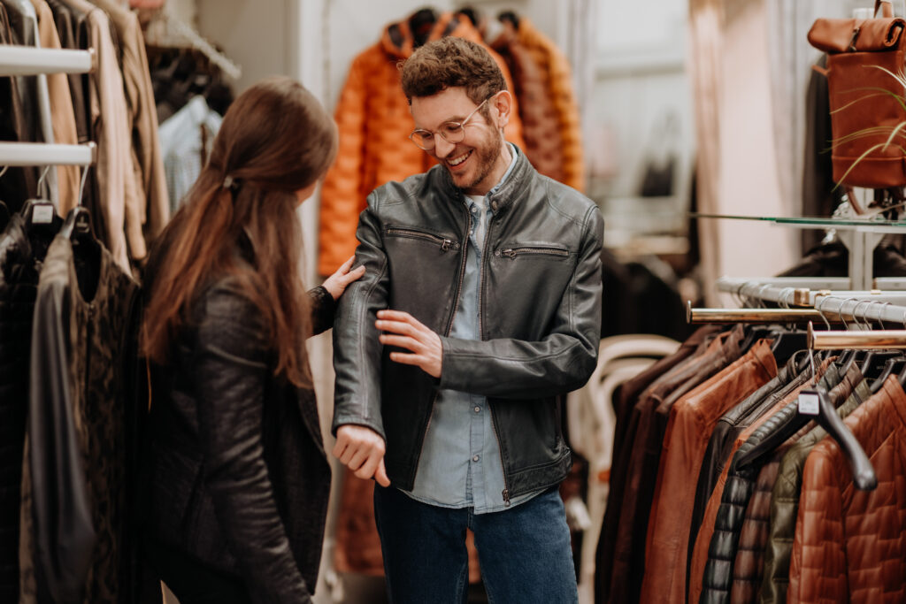 Mann probiert schwarze Lederjacke an, Verkäuferin berät ihn freundlich im Geschäft.