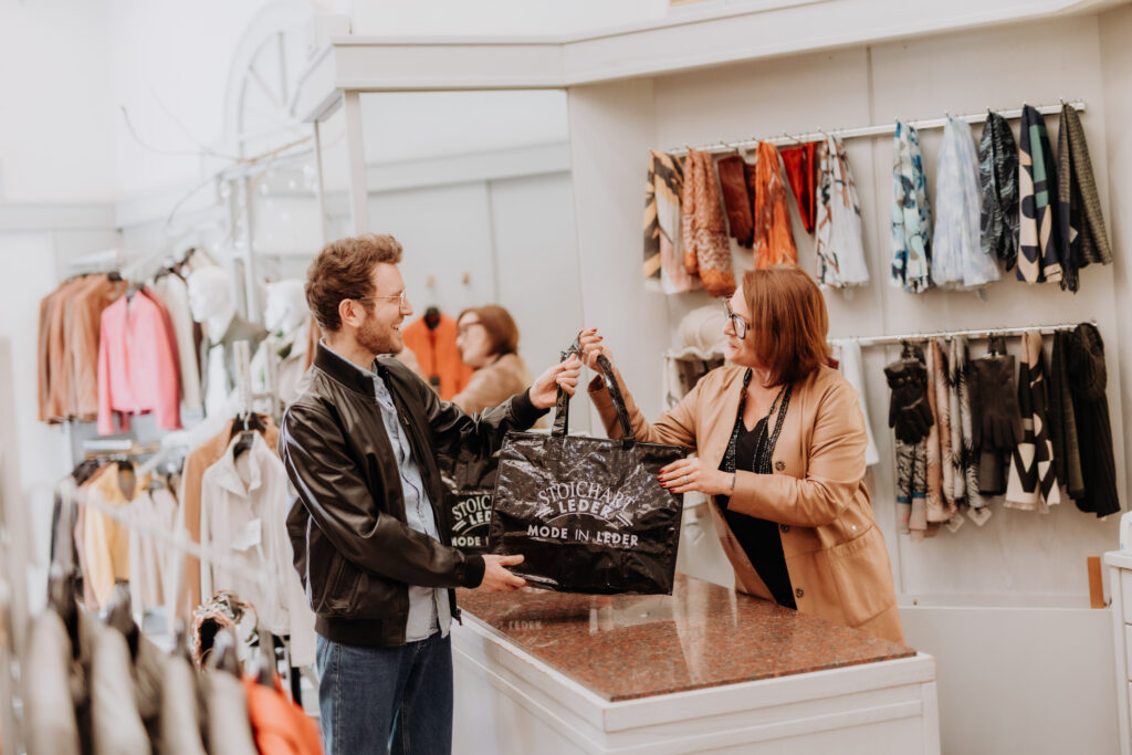 Ein Mann erhält eine Einkaufstasche mit der Aufschrift „Stoichart Leder – Mode in Leder“ von einer Verkäuferin im Geschäft.