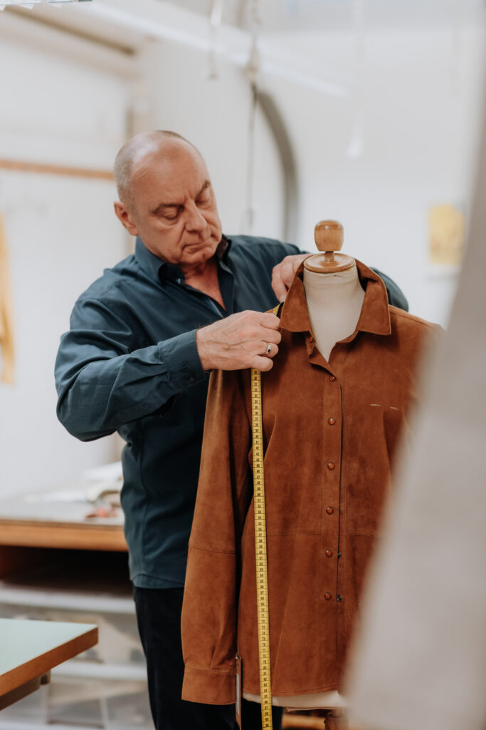 Ein Schneider misst mit einem Maßband ein braunes Lederhemd auf einer Schneiderpuppe im Atelier aus.
