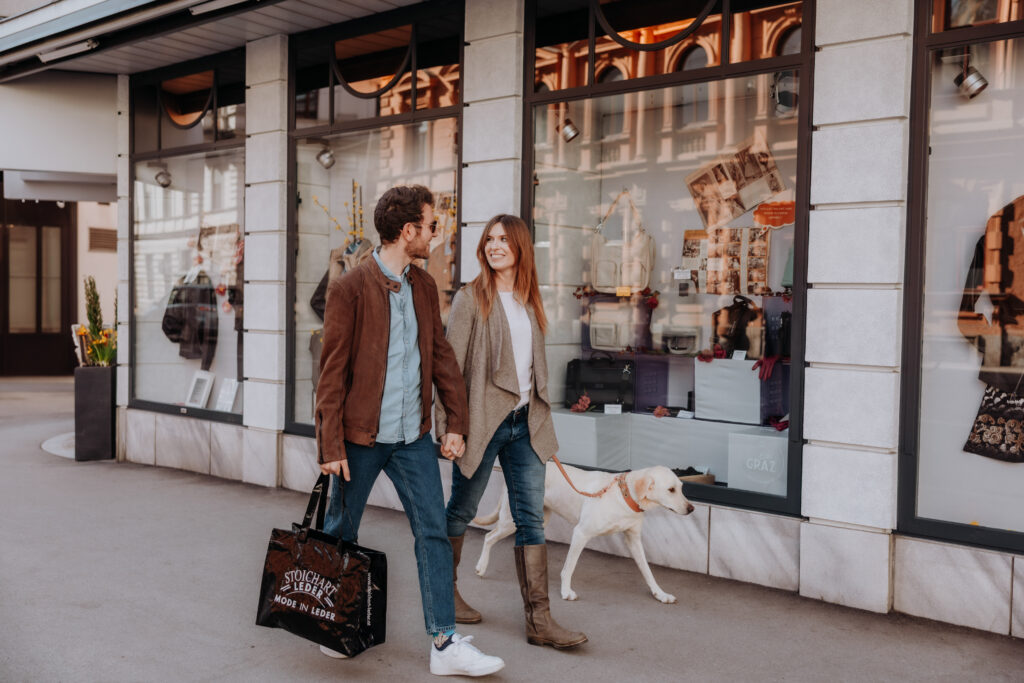 Ein Paar spaziert Hand in Hand mit Hund und Stoichart-Einkaufstasche vor einem Schaufenster in Graz entlang.