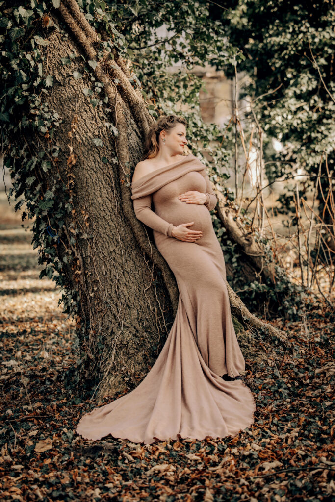 Schwangere Frau in langem beigefarbenem Kleid lehnt an einem alten Baum im herbstlichen Wald